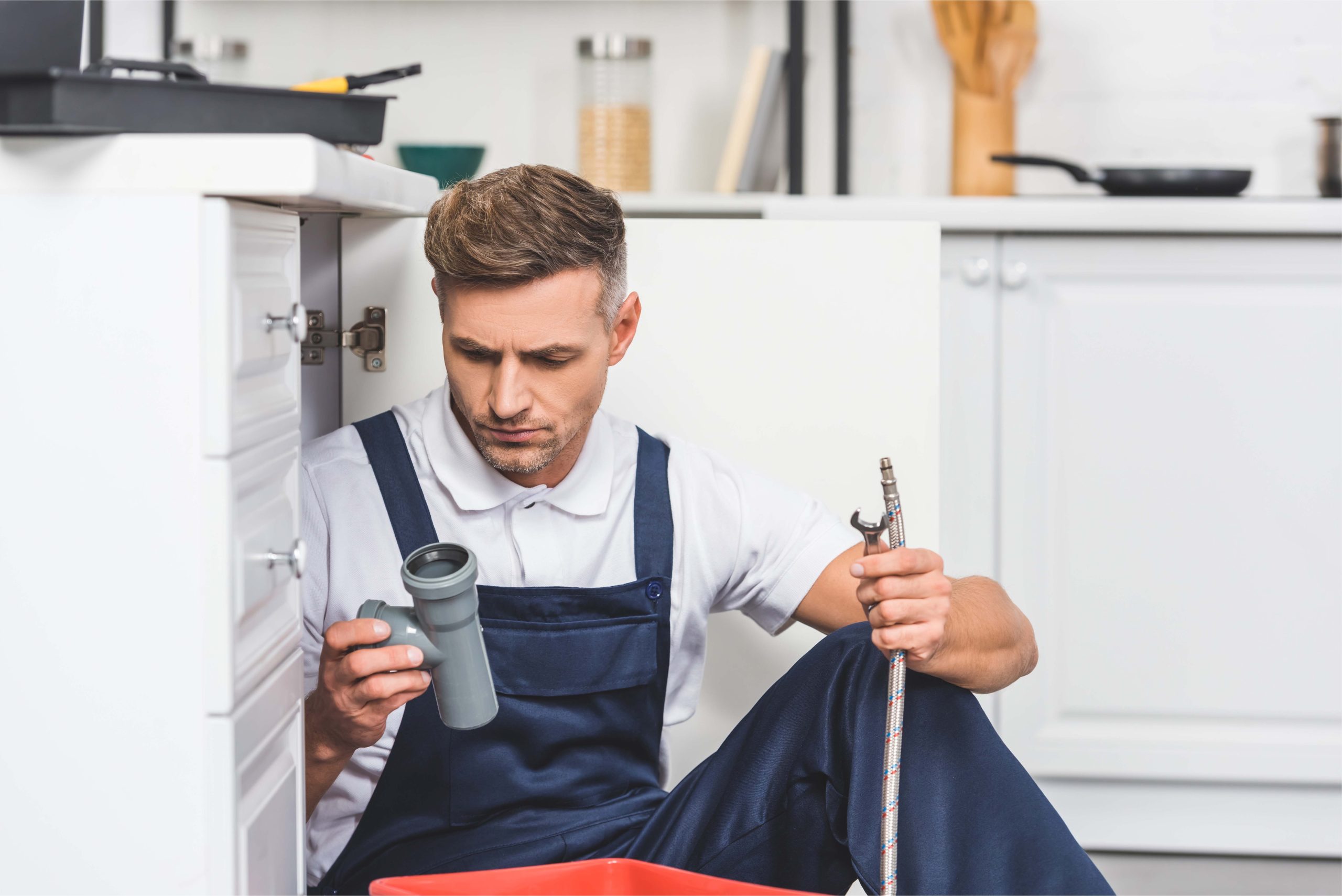 thoughtful adult repairman sitting under sink and holding pipes