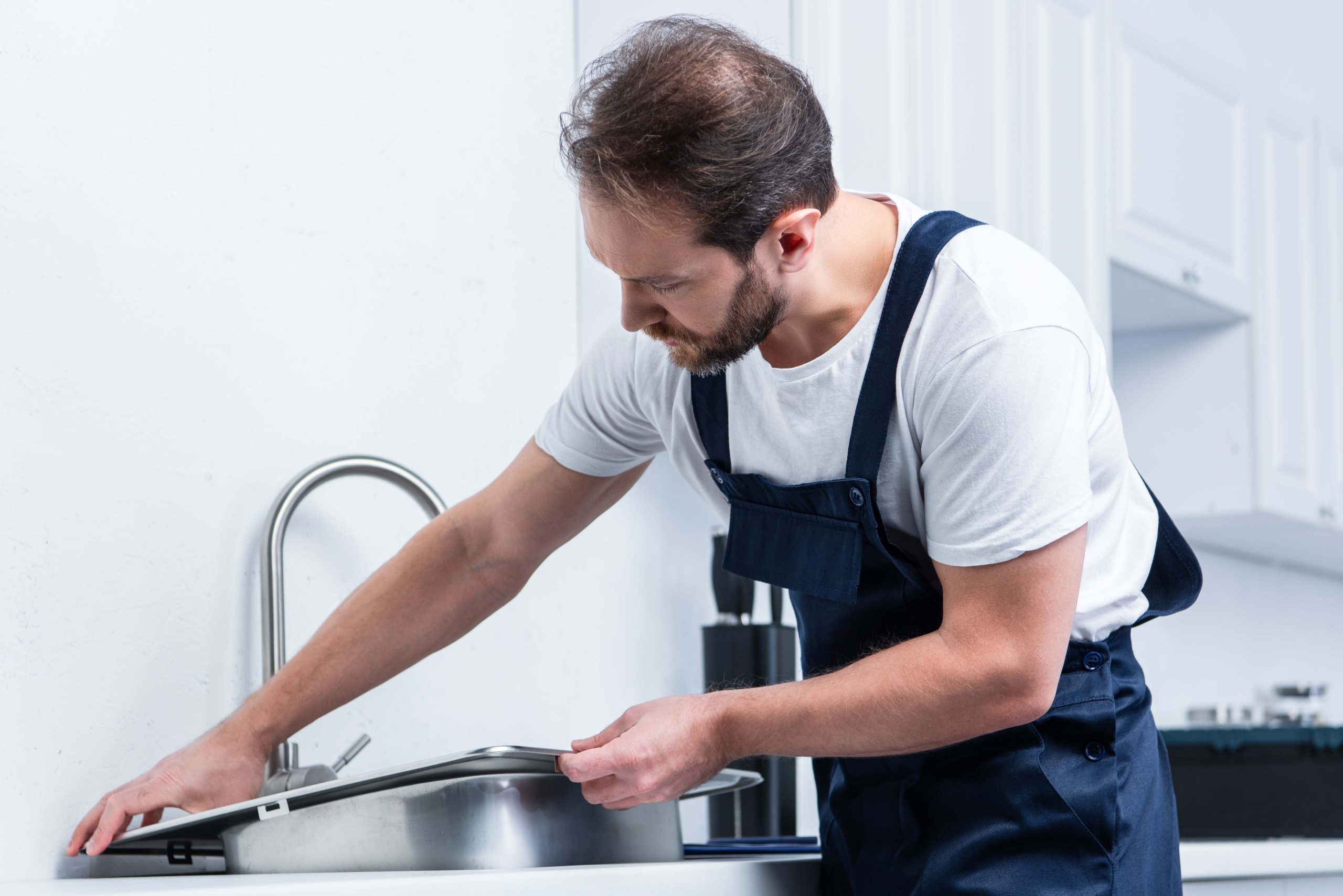 adult bearded repairman in working overall fixing sink in kitche